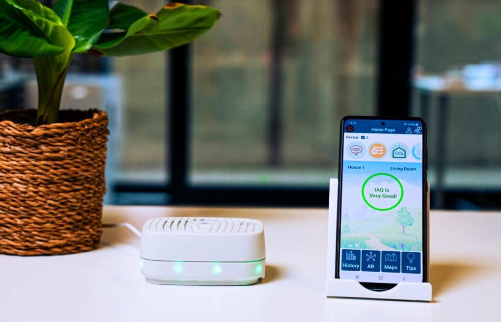 18-in-1 Indoor Air Quality Monitor with Dual Chips devices are on table with smart phone with it also flower plot are there with device 