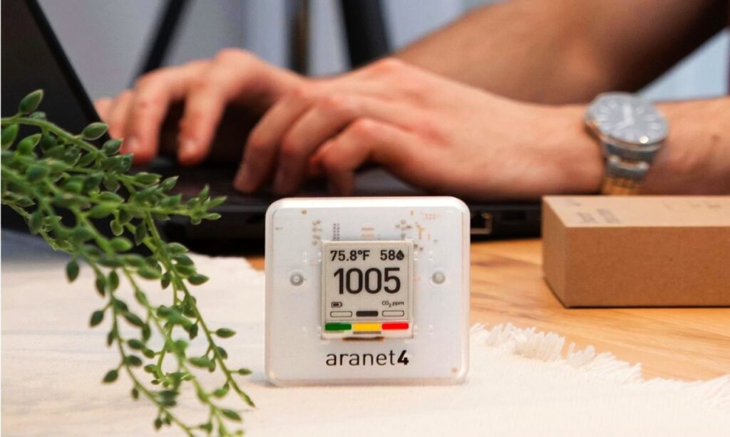SAF Aranet4 Home Wireless Indoor Air Quality Monitor device are laying on table a person using laptop 
