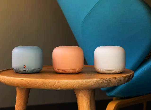there are three Google Nest Wifi devices are on brown colored table devices having grey white and orange colors 