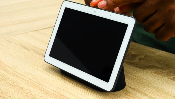 a person touch the Google Nest Hub Max device that laying on table its color is white and black