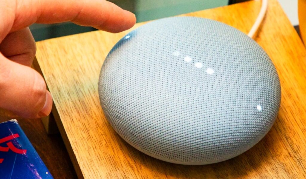 a person going to touch the Google Nest Mini device that laying on table its color is grey